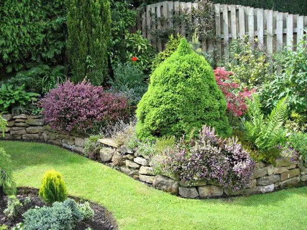 Raised natural stone garden bed with ornamental shrubs, purple bushes, evergreen trees, and manicured green lawn